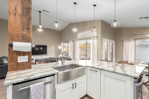 A modern kitchen with a granite countertop, stainless steel sink, and white cabinetry. There is a wooden accent wall, pendant lighting, and a flat-screen TV mounted on the wall.