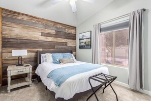A bedroom with a wooden headboard, a bed with white bedding and blue accents, a bedside table with a lamp, a window with curtains, and a picture on the wall.