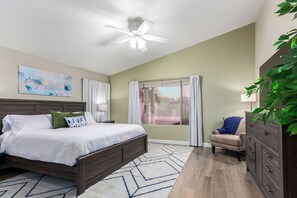 Une chambre à coucher avec un lit, un ventilateur de plafond, une fenêtre avec des rideaux, une chaise, une commode et un tableau accroché au mur.