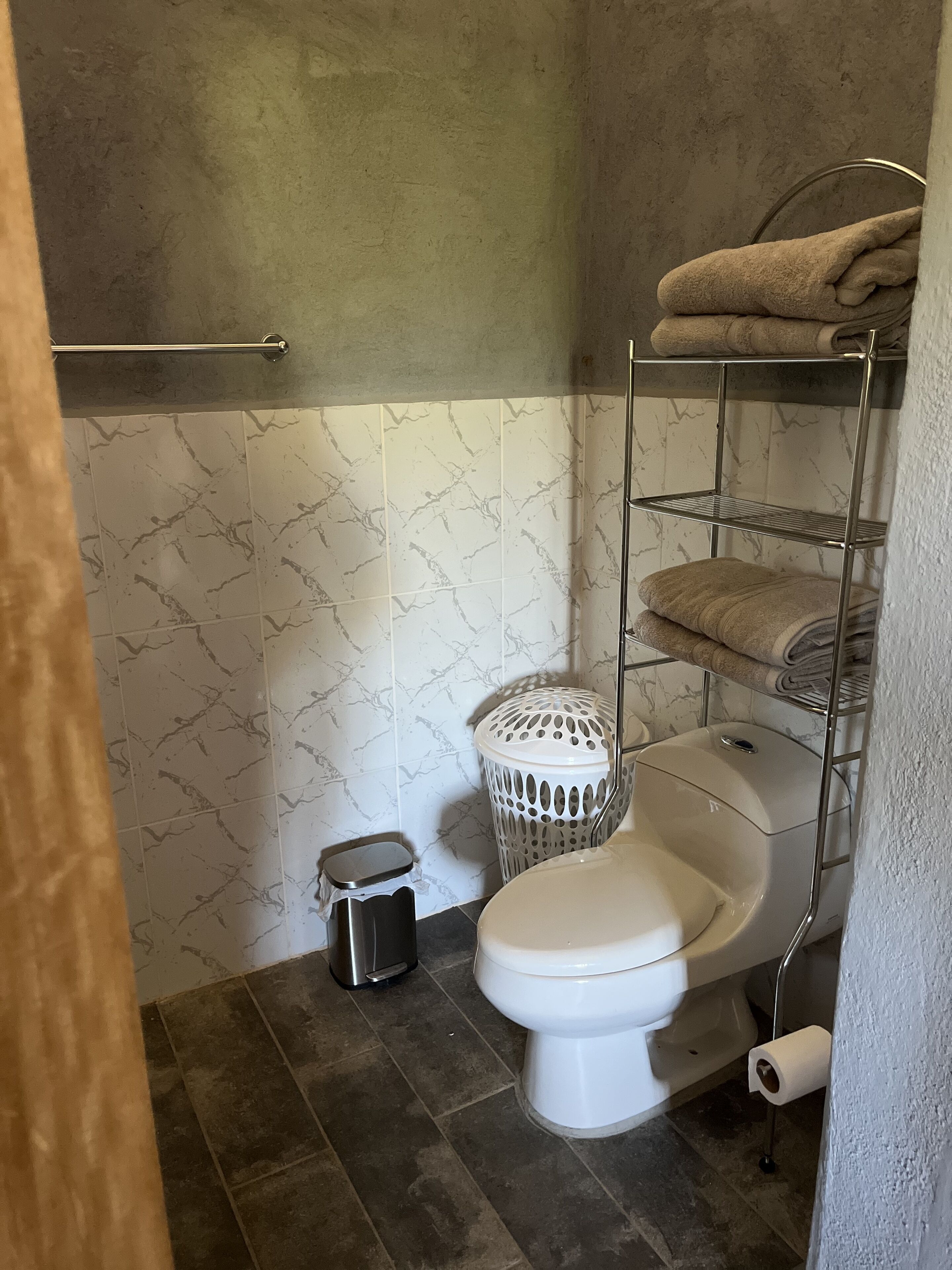 Ein Badezimmer mit Toilette, einem Metallregal mit Handtüchern, einem Mülleimer und einem Toilettenpapierhalter.