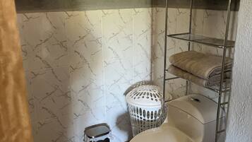 Ein Badezimmer mit Toilette, einem Metallregal mit Handtüchern, einem Mülleimer und einem Toilettenpapierhalter.