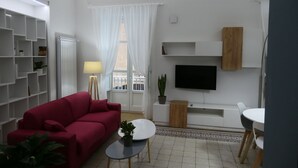 A modern living room with a red sofa, a white coffee table, a TV, and a dining area with a white table and chairs. - Opera Suites - Tosca (Palermo)