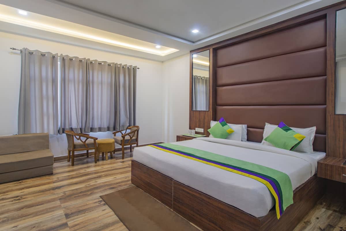 Habitación de hotel con cama de madera, mesita de noche de madera, una silla, un pequeño sofá y una ventana con cortinas.