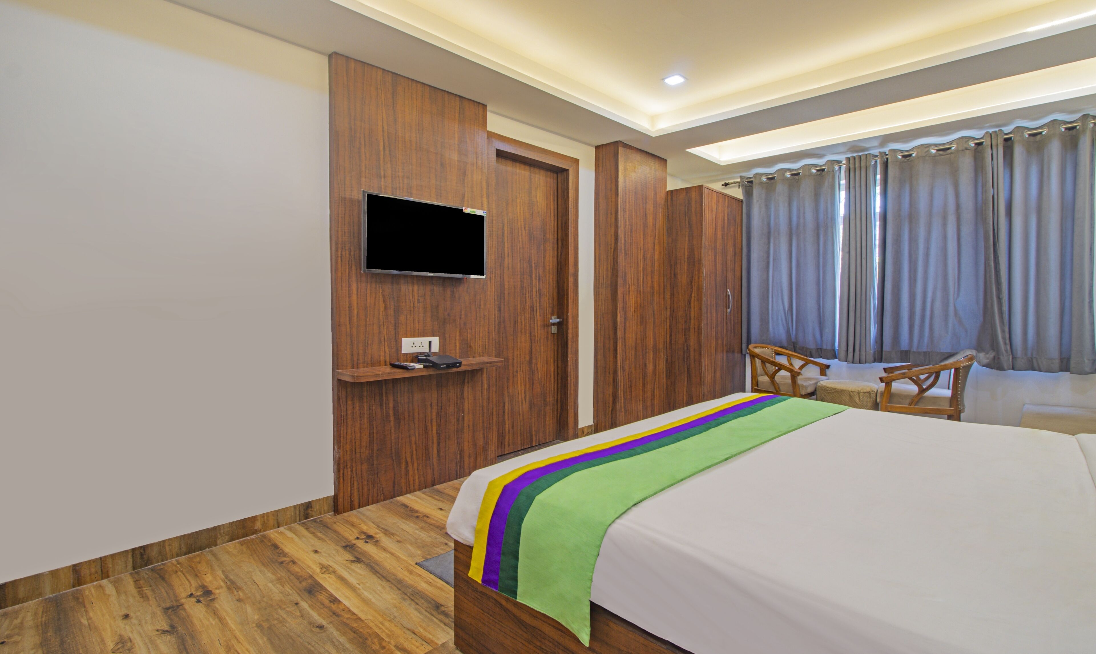 Habitación de hotel con un armario de madera, un televisor de pantalla plana montado en la pared, una cama con una manta colorida, una silla de madera y una ventana con cortinas.