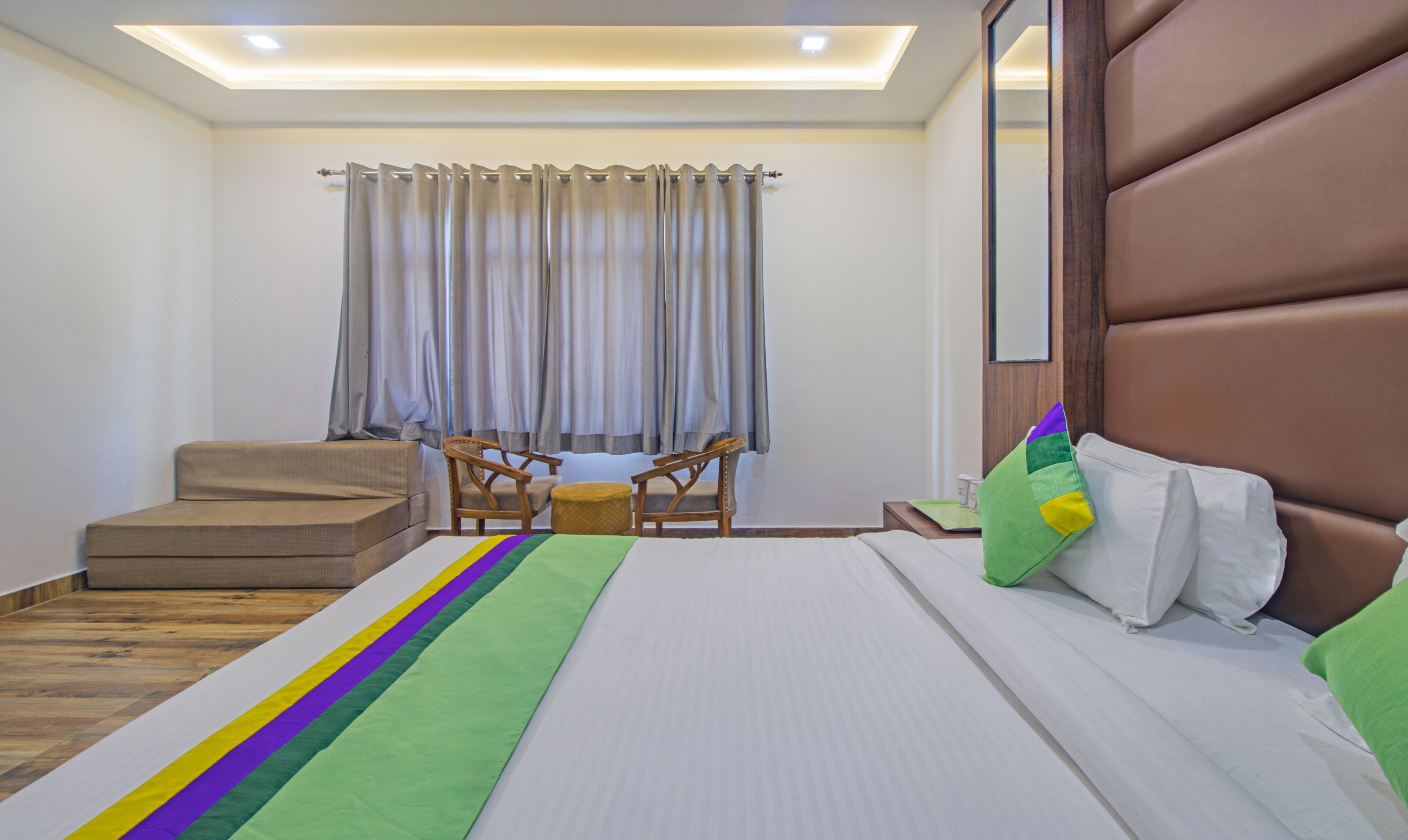 Habitación de hotel con cama, banco de madera, dos sillas, una mesita y una ventana con cortinas.