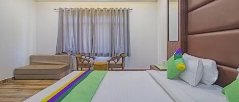 Habitación de hotel con cama, banco de madera, dos sillas, una mesita y una ventana con cortinas.
