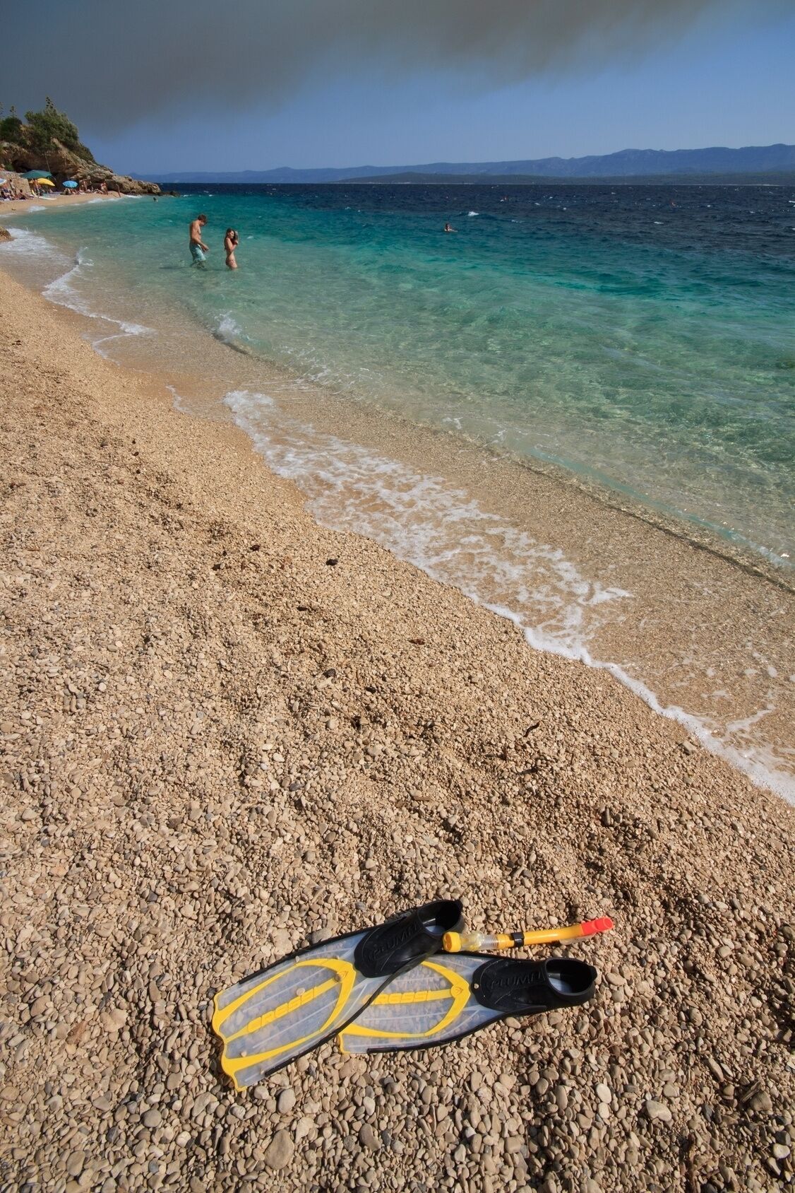 A pebble beach with clear water, a pair of yellow and black flippers, and a distant person swimming.