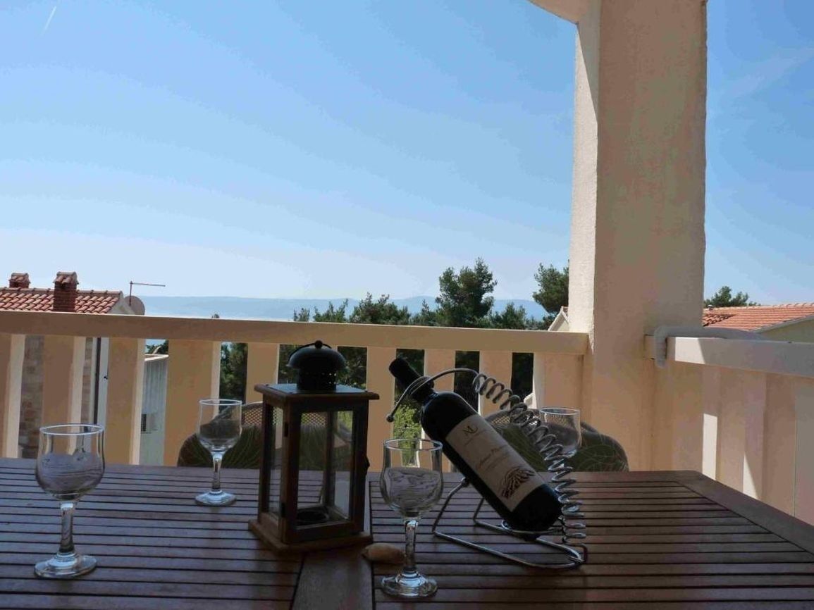 A wooden table with wine glasses, a bottle, and a lantern on a balcony with a view of trees and a building in the background.