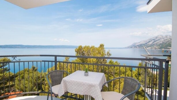 A balcony with a table and two chairs, overlooking a body of water and greenery.