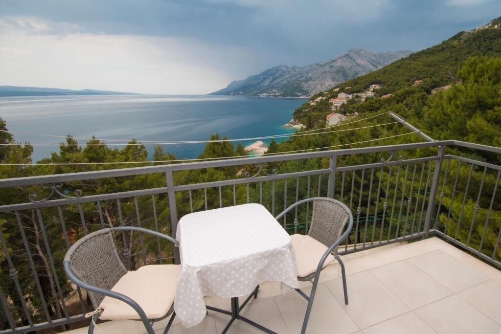 A balcony with a table and two chairs, overlooking a coastal landscape with mountains and a body of water.
