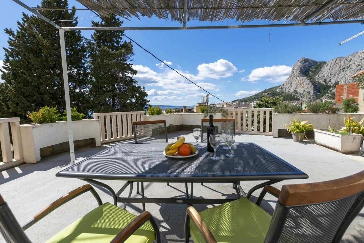 A rooftop terrace with a table set for two, featuring a bottle and glasses, and a plate of fruit.