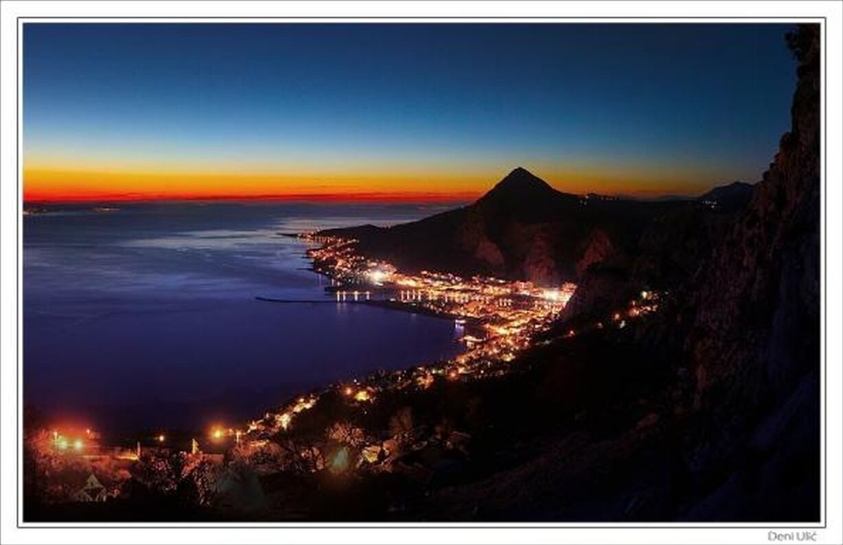 A coastal town at dusk with a mountain in the background and a pier extending into the water.