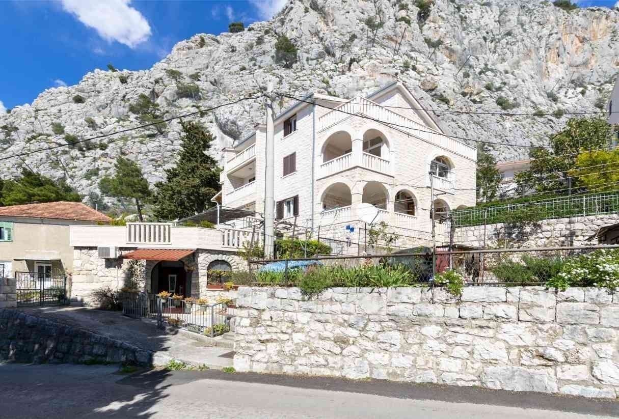 A multi-story building with a stone facade, arched windows, and a balcony. It is situated in a mountainous area with rocky terrain in the background.