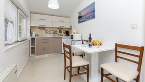A compact kitchen with a dining area, featuring a white table, two wooden chairs, a white pendant light, and a painting of a coastal scene. - Apartment in Omiš with Seaview, Balcony, Air condition, WIFI (5129-5) (Omiš)