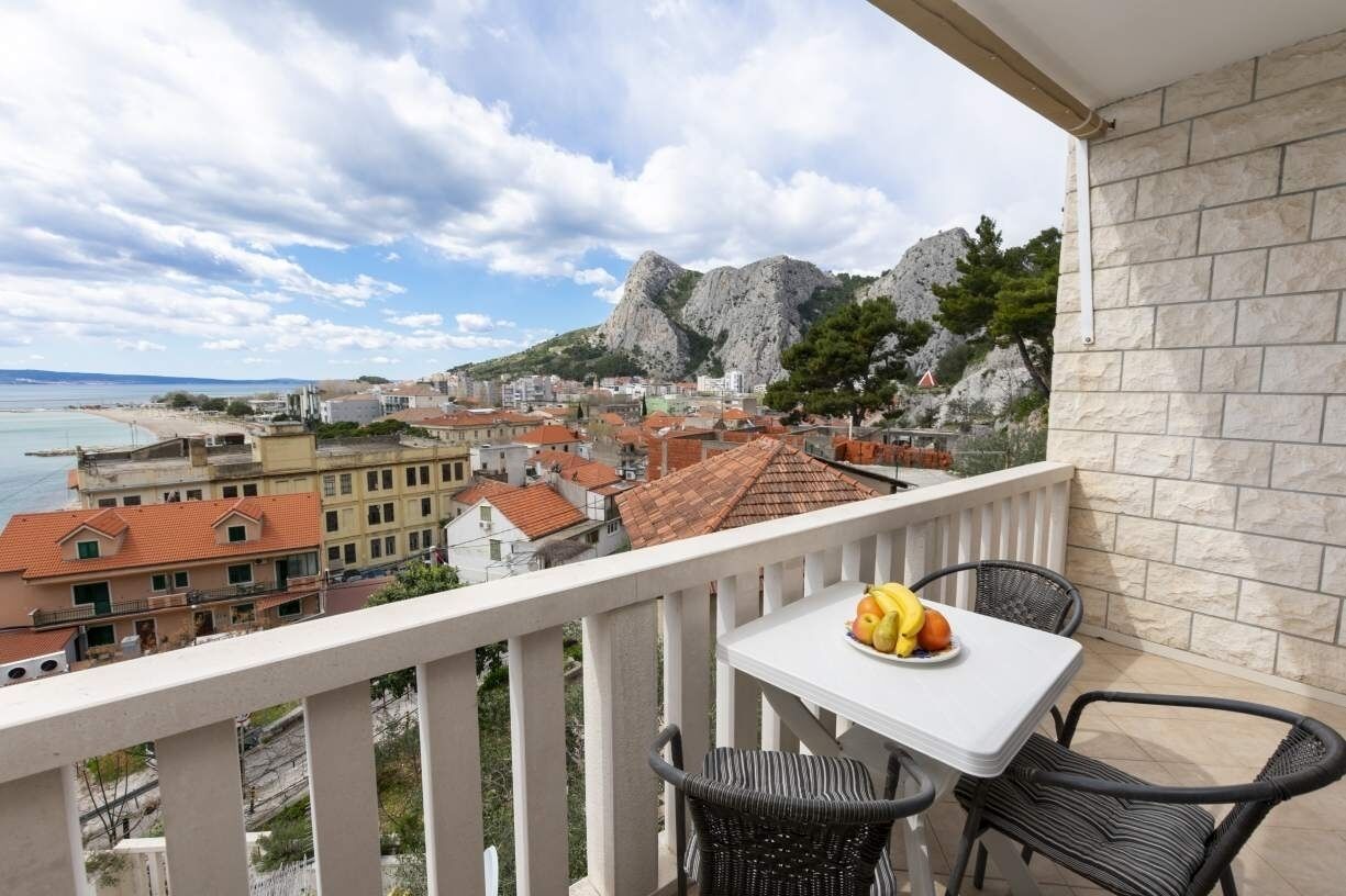 A balcony with a table and chairs, overlooking a coastal town with buildings and mountains.