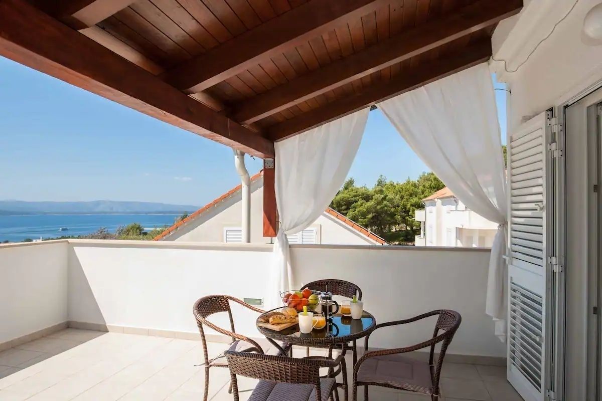 Ein Balkon mit Korbstühlen, einem Tisch mit Frühstücksgebäck und Blick auf das Meer und die Berge.