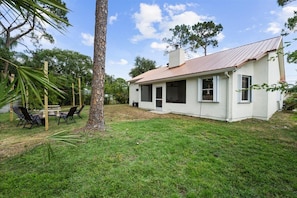 A single-story house with a white exterior and a red roof, surrounded by a grassy yard and trees.