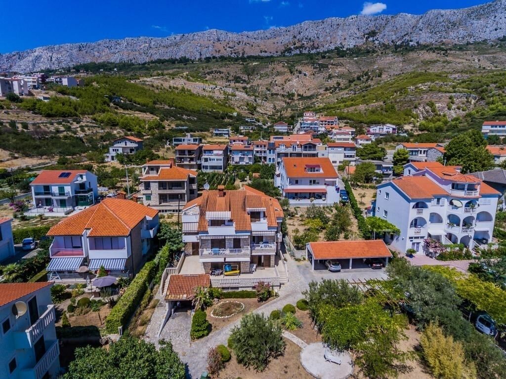A residential area with houses featuring red-tiled roofs, surrounded by greenery and rocky hills in the background.
