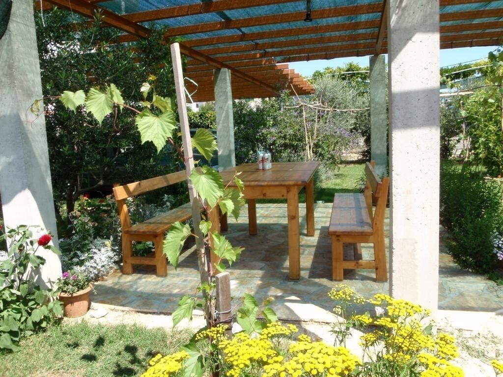 A covered outdoor seating area with a wooden table and benches, surrounded by potted plants and flowers.