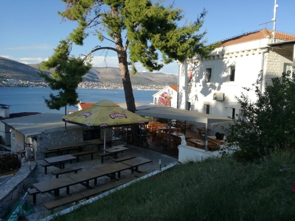 An outdoor seating area with tables and a yellow umbrella, overlooking a body of water and a town in the distance.