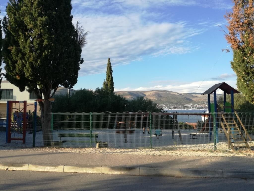 Ein eingezäunter Spielplatz mit Spielgeräten, einer kleinen Schutzhütte und Blick auf einen See sowie Hügel im Hintergrund.