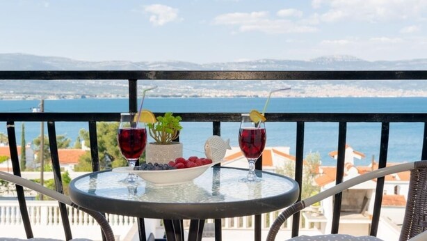 A balcony with a table set for two, featuring a bowl of fruit and two glasses of red wine, overlooking a coastal view.