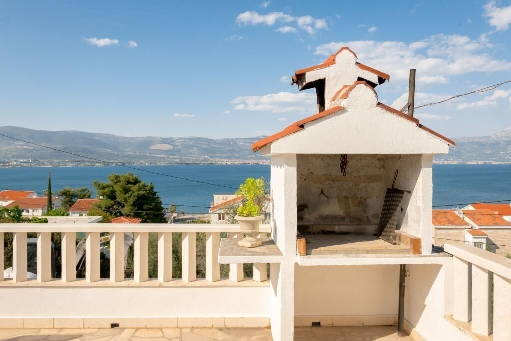 A white building with a red roof, a balcony with a railing, and a view of the sea and distant hills.