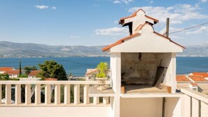 A white building with a red roof, a balcony with a railing, and a view of the sea and distant hills.