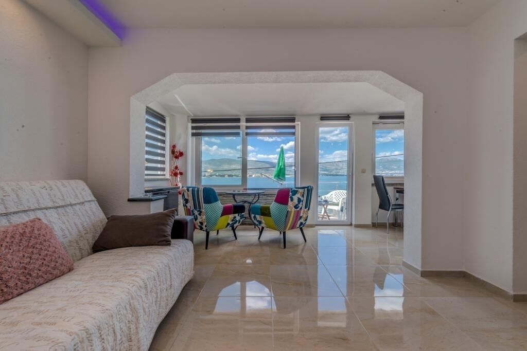 A living room with a sofa, colorful chairs, a table, and a view of the sea through large windows.