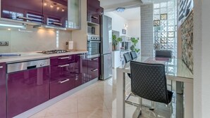 A modern kitchen with purple cabinets, stainless steel appliances, and a glass dining table with a black chair.