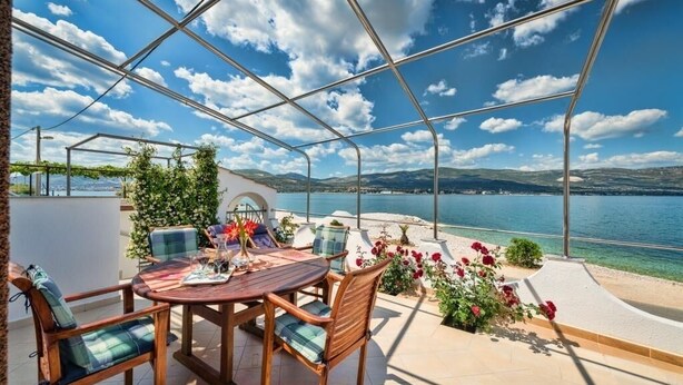 A covered outdoor seating area with a table and chairs, overlooking a body of water and hills.