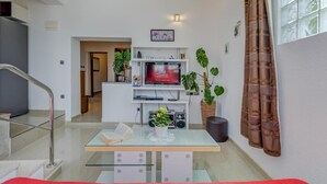 A modern living room with a glass coffee table, a flat-screen TV, and potted plants.