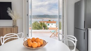 A dining area with a table, chairs, a basket of oranges, and a view of a coastal landscape through large windows.