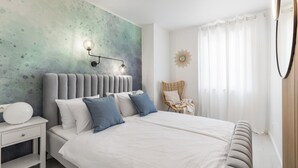 A bedroom with a large bed, a nightstand, a wall-mounted lamp, a chair, and a window with curtains.