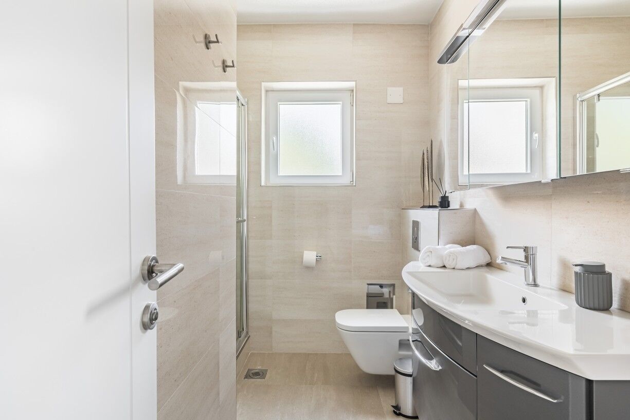 Un baño moderno con inodoro, lavamanos y ducha. Hay dos ventanas que dejan entrar luz natural.