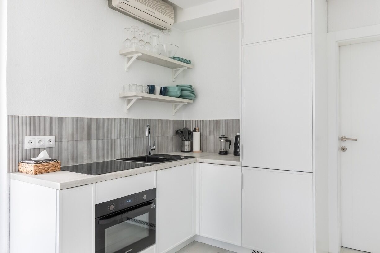 A modern kitchen with white cabinets, a black countertop, and a built-in oven. There are white shelves above the sink holding dishes and glasses.