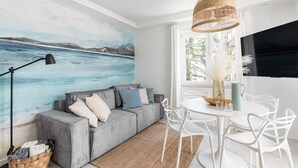 A modern living room with a grey sofa, white chairs, a round table, a large painting of a seascape, a wicker pendant light, and a black TV on a white wall.