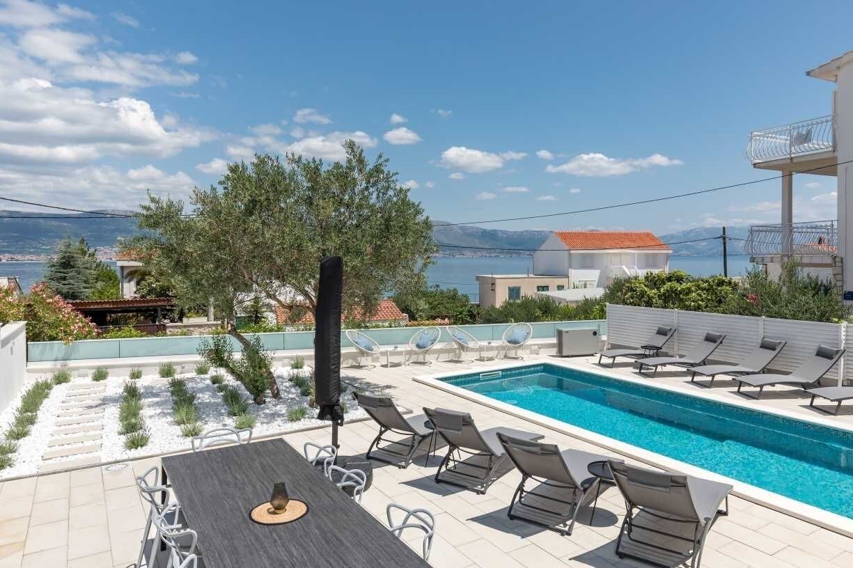 A rooftop pool area with lounge chairs, a table, and a view of the sea and mountains.