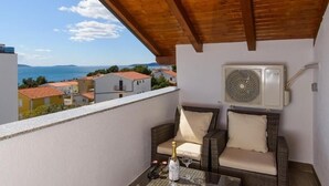 Un balcon avec vue sur la mer, un canapé en osier, une table avec une bouteille et des verres, et un appareil de climatisation.