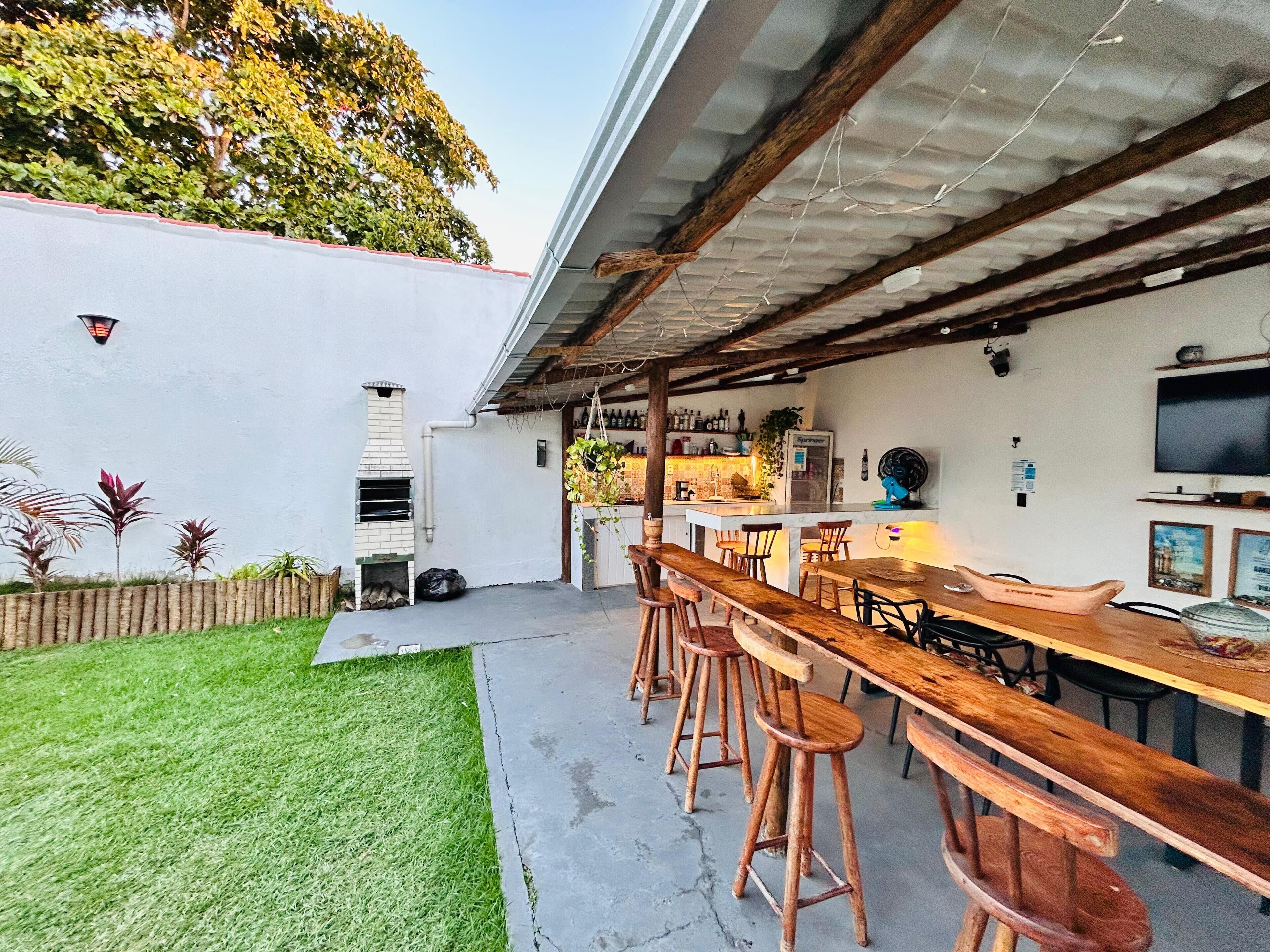 Eine Terrasse im Freien mit einem langen Holztisch, Barhocker, einem Backofen aus Ziegelsteinen und einem an der Wand befestigten Fernseher.