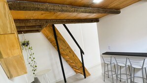 A modern loft-style room with a high ceiling, exposed wooden beams, a grey sofa, a dining table with chairs, and a staircase. - Pousada Atenas (Porto Seguro)