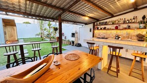 An outdoor dining area with a wooden table and chairs, a bar with various bottles, and a view of a garden. - Pousada Atenas (Porto Seguro)