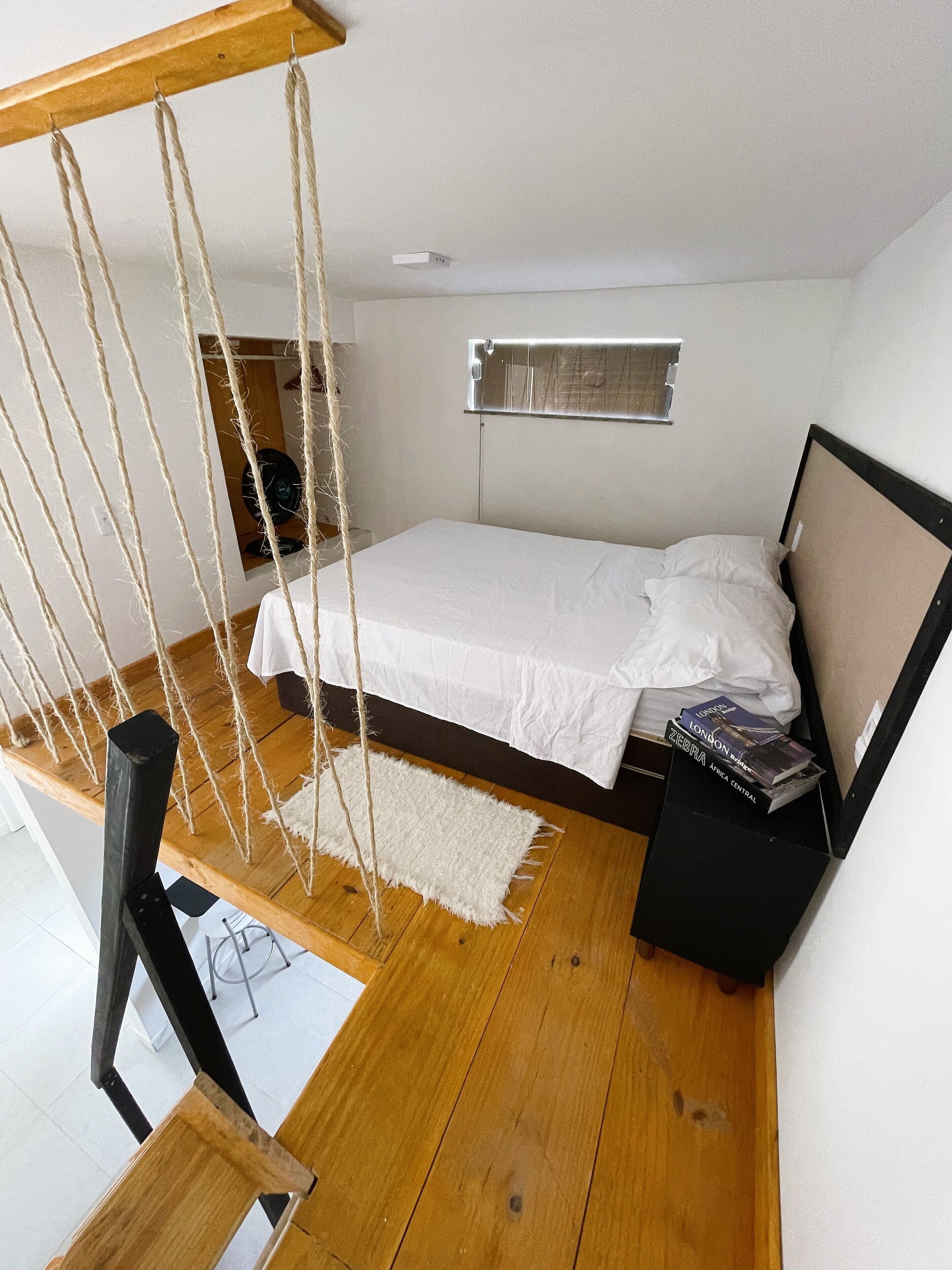 A bedroom with a bed, a hanging chair, a small rug, and a bedside table with books.