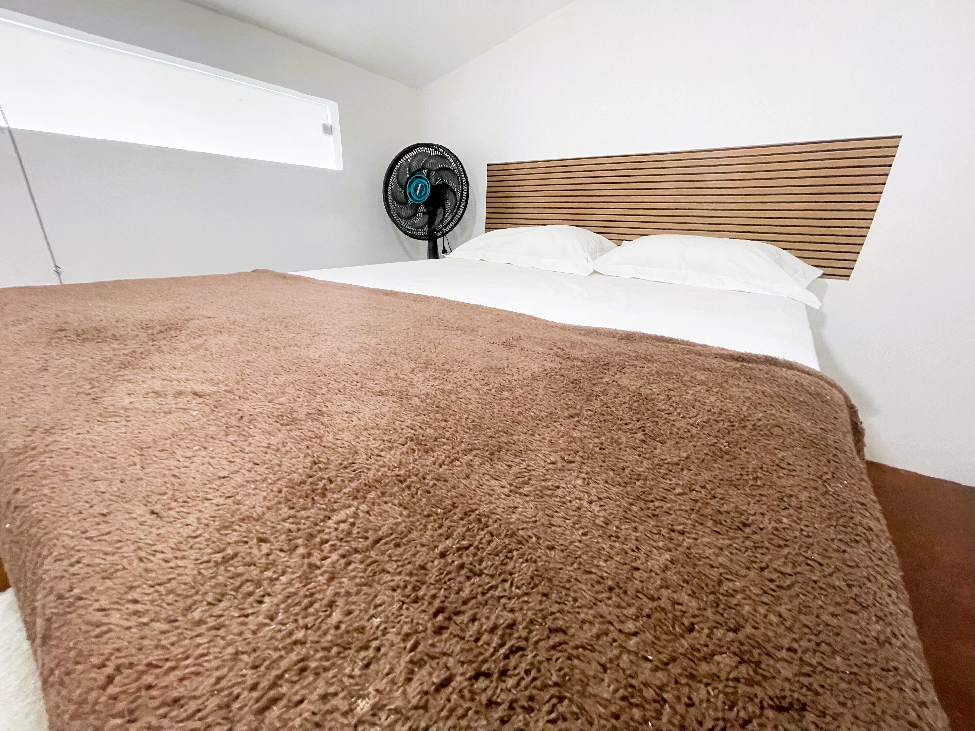A bedroom with a large bed, a wooden headboard, a standing fan, and a window with blinds.