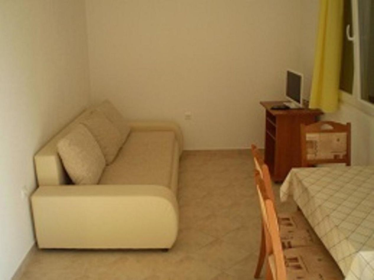 Quarto pequeno e compacto com sofá, cama, mesa de cabeceira de madeira com televisão e janela com cortinas.