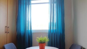Ein kleiner Raum mit einem runden Tisch, zwei Stühlen, einer Topfpflanze und einem Fenster mit blauen Vorhängen.