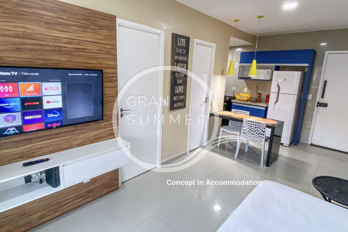 Ett modernt hotellrum med en platt-TV, en vägg med träpanel som ger rummet karaktär, ett köksutrymme med kylskåp och ett matbord med stolar.