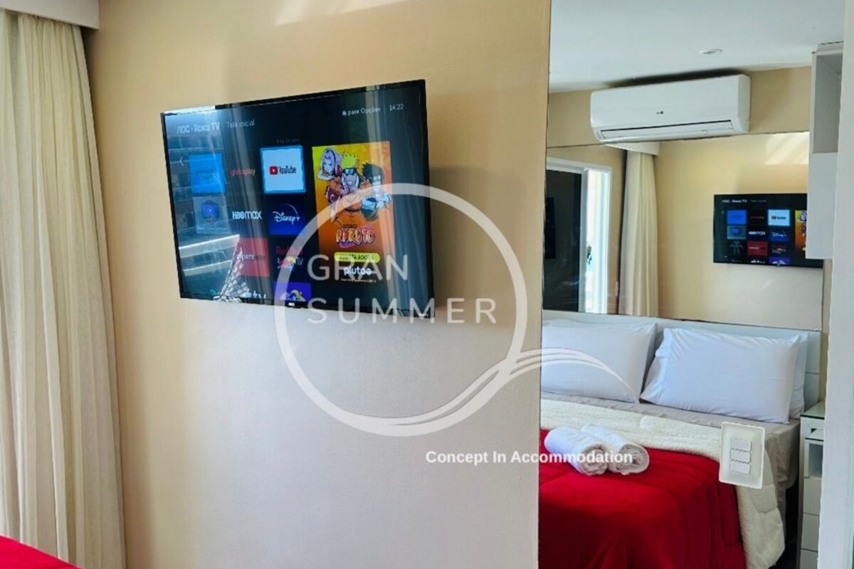 A hotel room with a bed, a TV mounted on the wall, and an air conditioning unit.