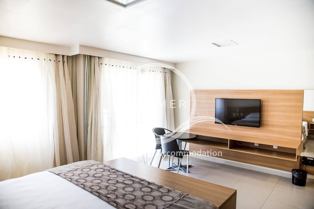 Ett modernt hotellrum med en säng, en tv som är monterad på en träpanel, ett skrivbord med en stol och ett stort fönster med gardiner.