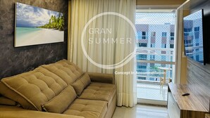 Una sala de estar moderna con un sofá, un cuadro de una playa, un ventanal con cortinas y un televisor sobre un mueble de madera.